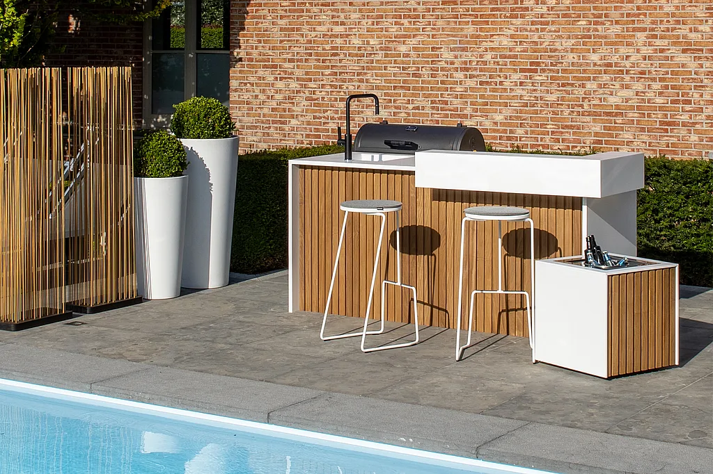 Witte buitenkeuken met houten latten naast een zwembad, met twee witte tiki barkrukken, hoge plantenbakken en een koperkleurige Sticks-scherm voor een bakstenen huis.