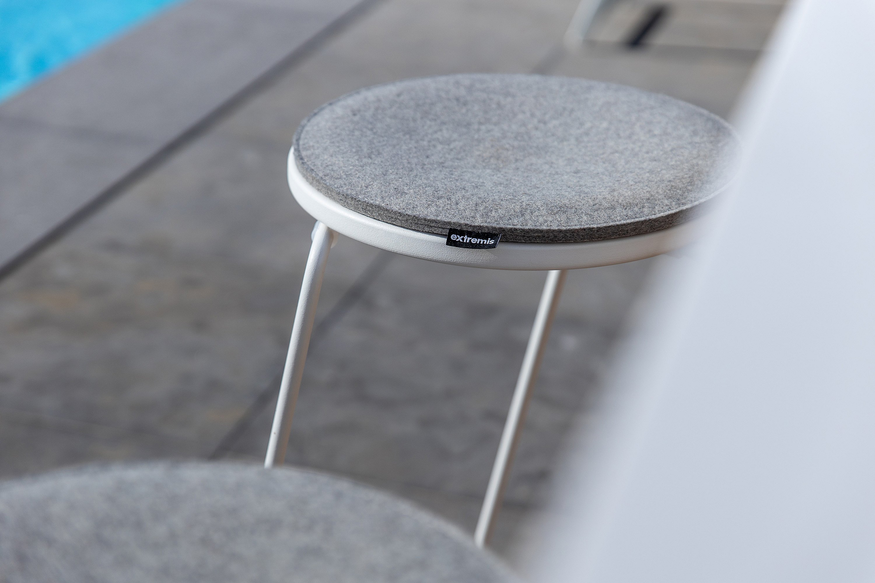 Close-up of a white barstool with a round grey upholstered seat on a stone terrace beside the pool.