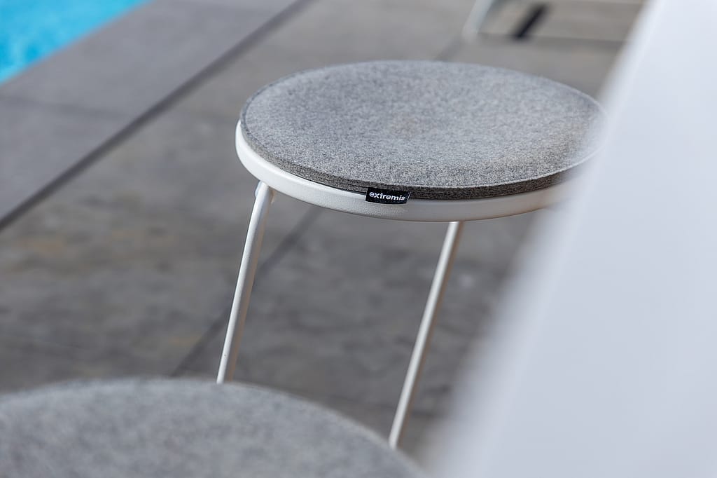 Close-up of a white barstool with a round grey upholstered seat on a stone terrace beside the pool.