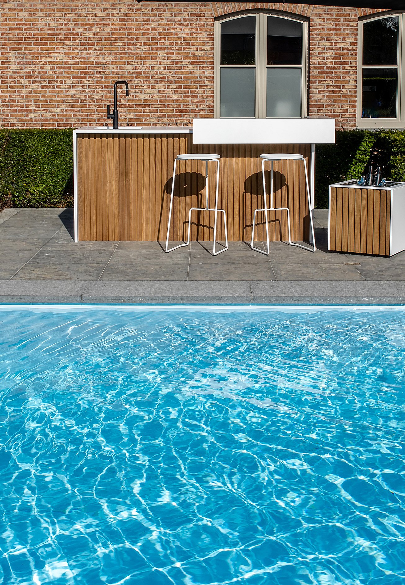 Front view of a white and wood outdoor kitchen with two white barstools beside a bright blue swimming pool, set against a brick house.