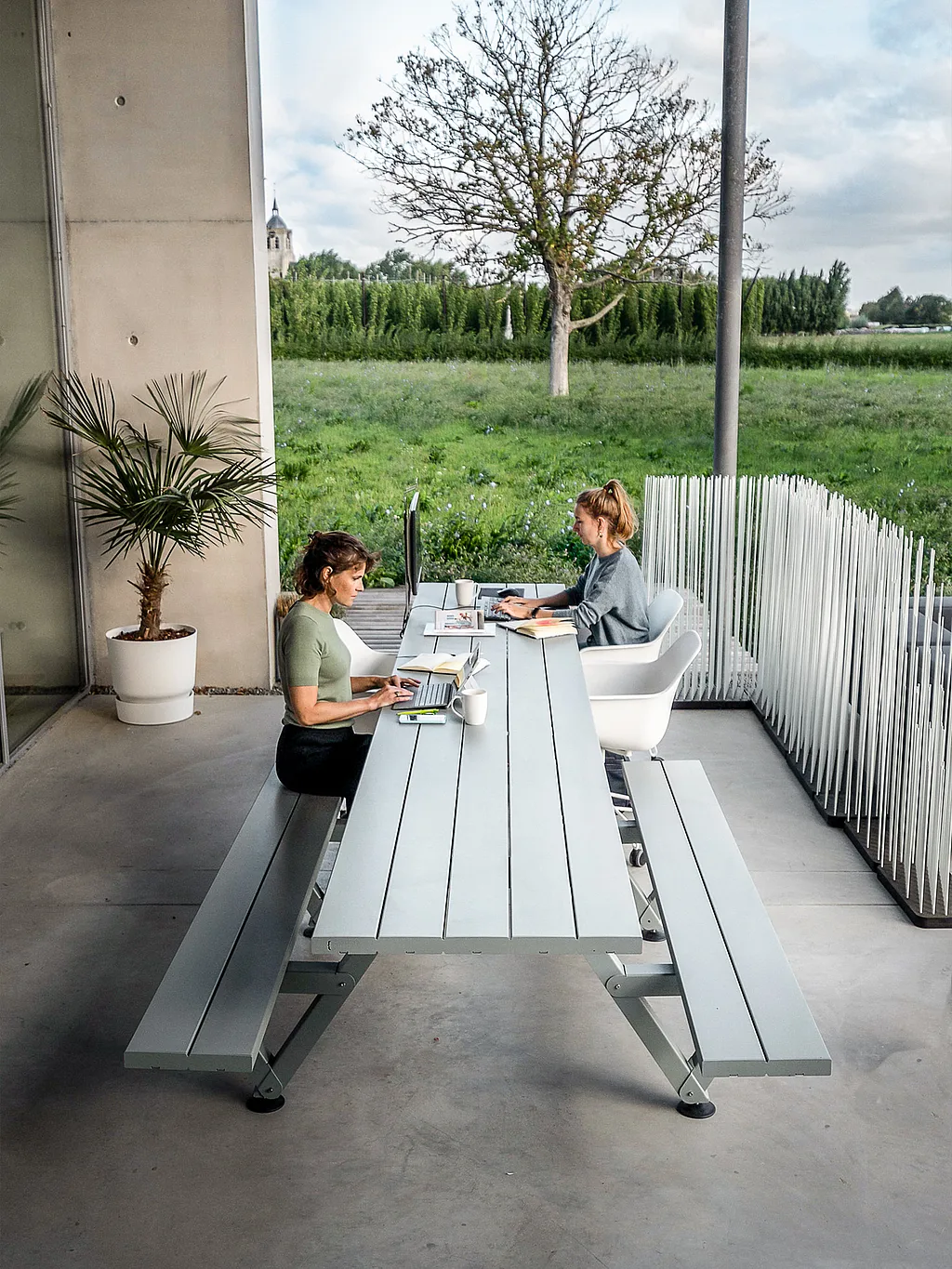 Twee collega's werken buiten aan een lange grijze jachthaventafel op een overdekt terras, met laptops, een monitor en open groene velden daarbuiten.