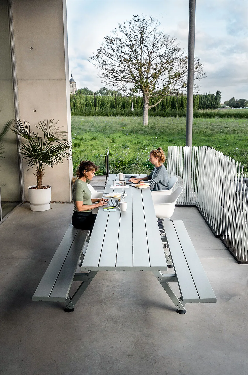 Two colleagues working outdoors at a long grey marina table on a covered terrace, with laptops, a monitor, and open green fields beyond.