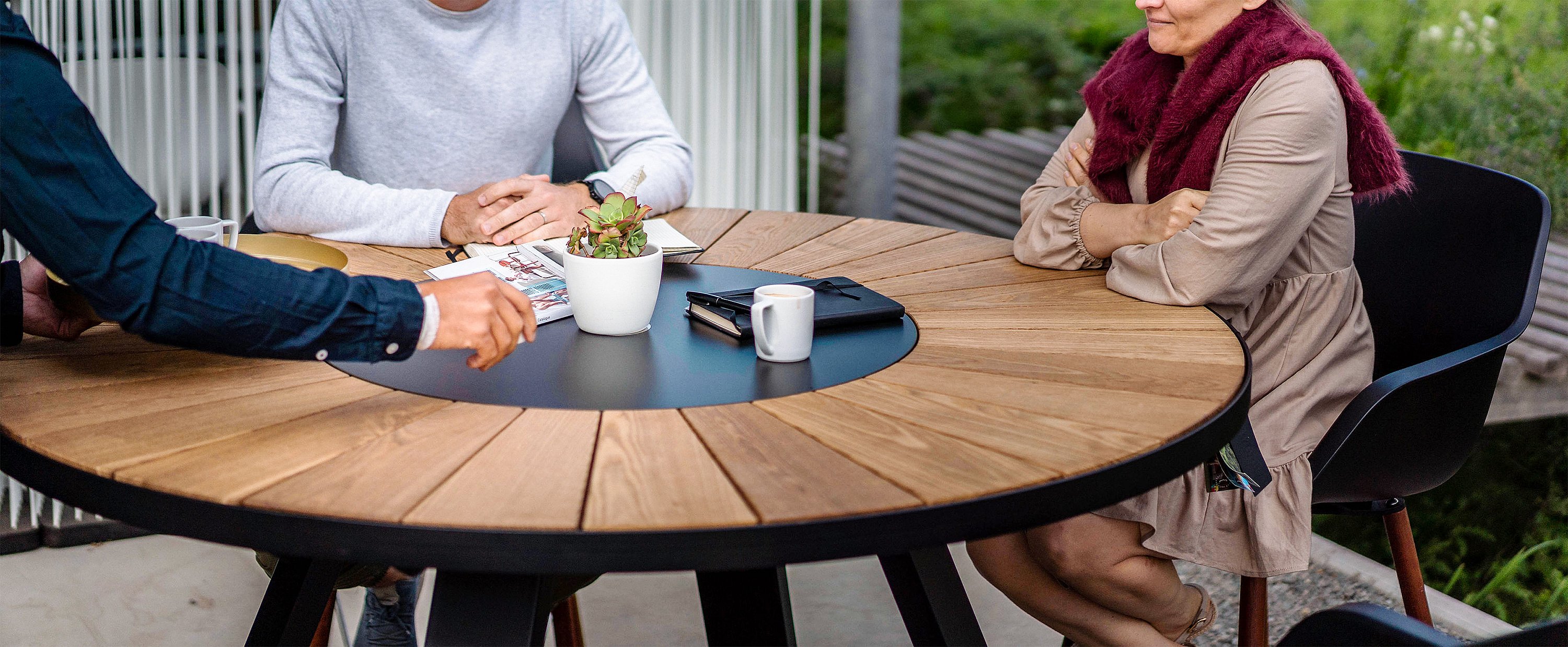 Vue rapprochée de trois collègues assis autour d’une table de réunion ronde en bois avec une plante, des cahiers et des tasses à café.