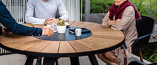 Vue rapprochée de trois collègues assis autour d’une table de réunion ronde en bois avec une plante, des cahiers et des tasses à café.