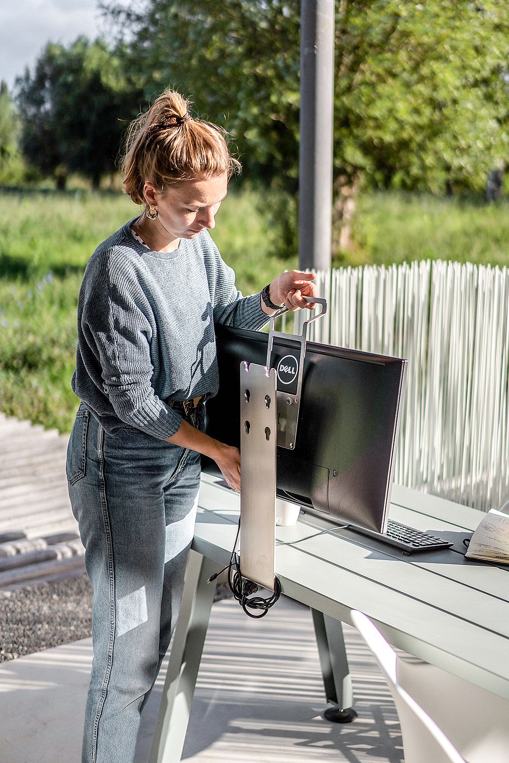 Une femme place un support d’écran sur une longue table de marina extérieure avec un écran, un clavier et un cahier en plein soleil.