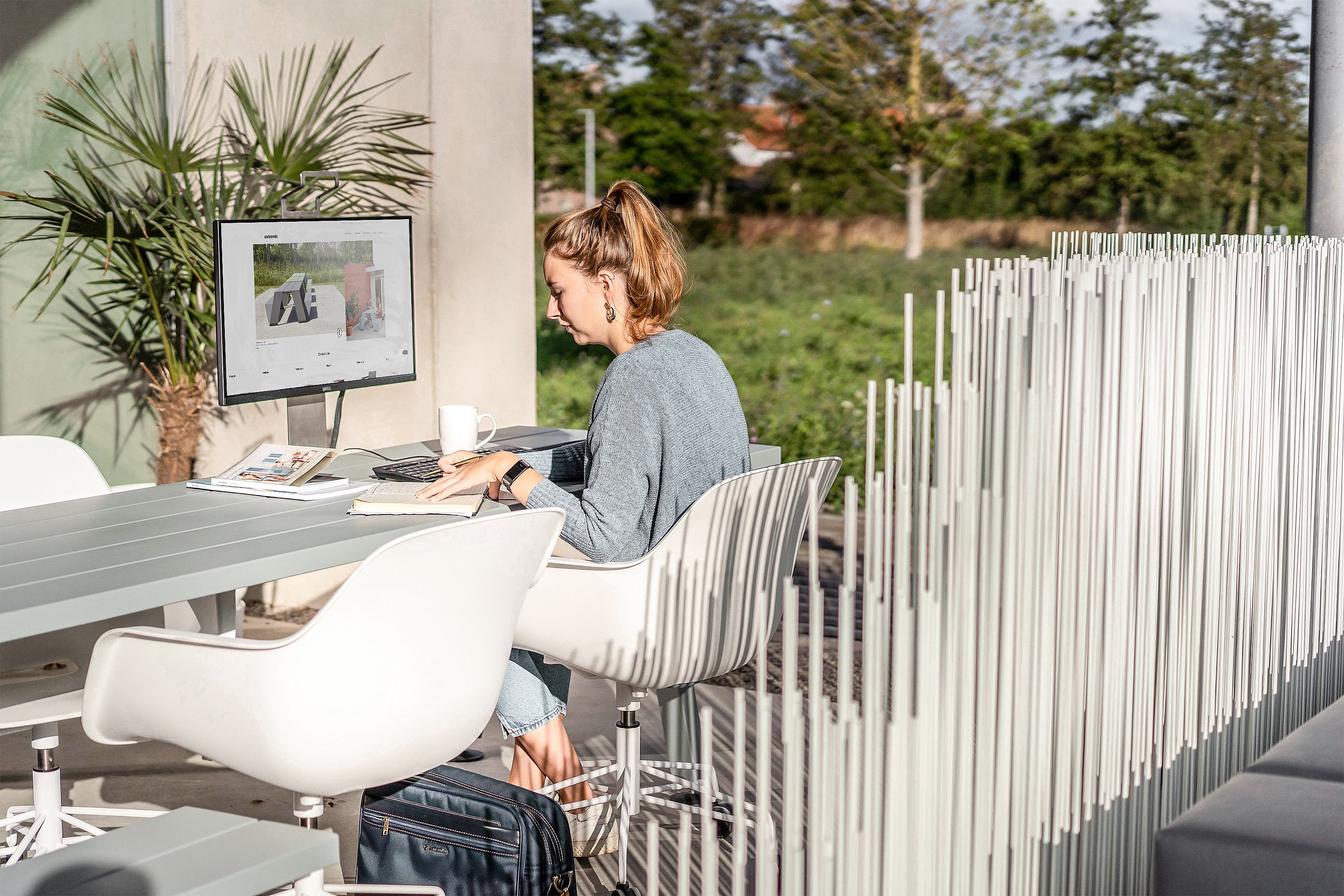 A woman works at a grey Marina outdoor table with a monitor, notebook and coffee mug beside a white vertical divider screen on a sunny terrace.