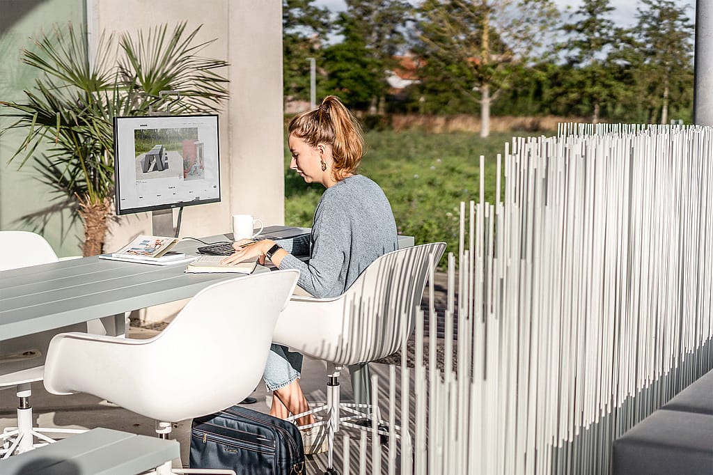 A woman works at a grey Marina outdoor table with a monitor, notebook and coffee mug beside a white vertical divider screen on a sunny terrace.