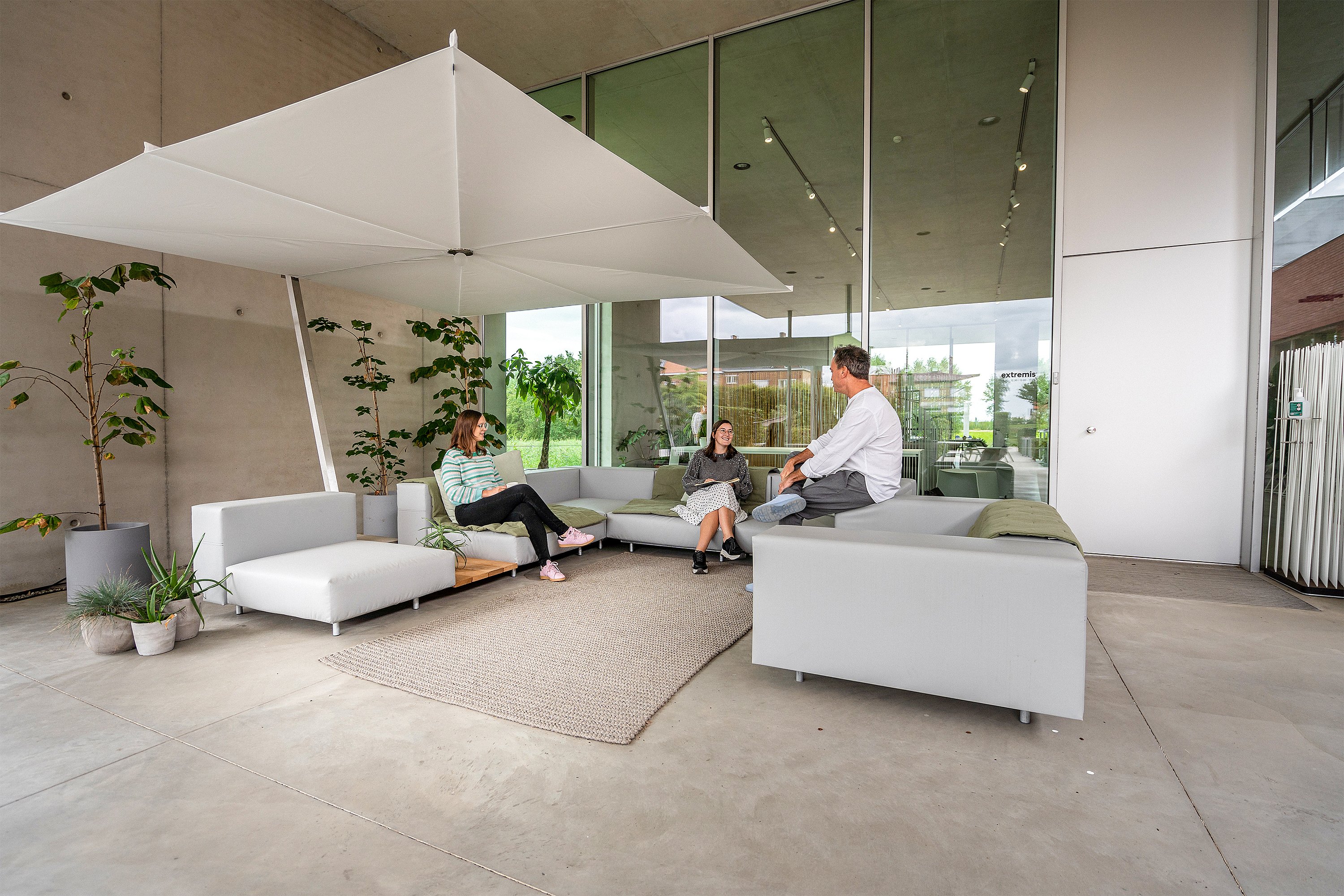 Large vue d’un walrus extérieur sous un grand parasol blanc, avec des canapés gris disposés autour d’un tapis sur une terrasse couverte.