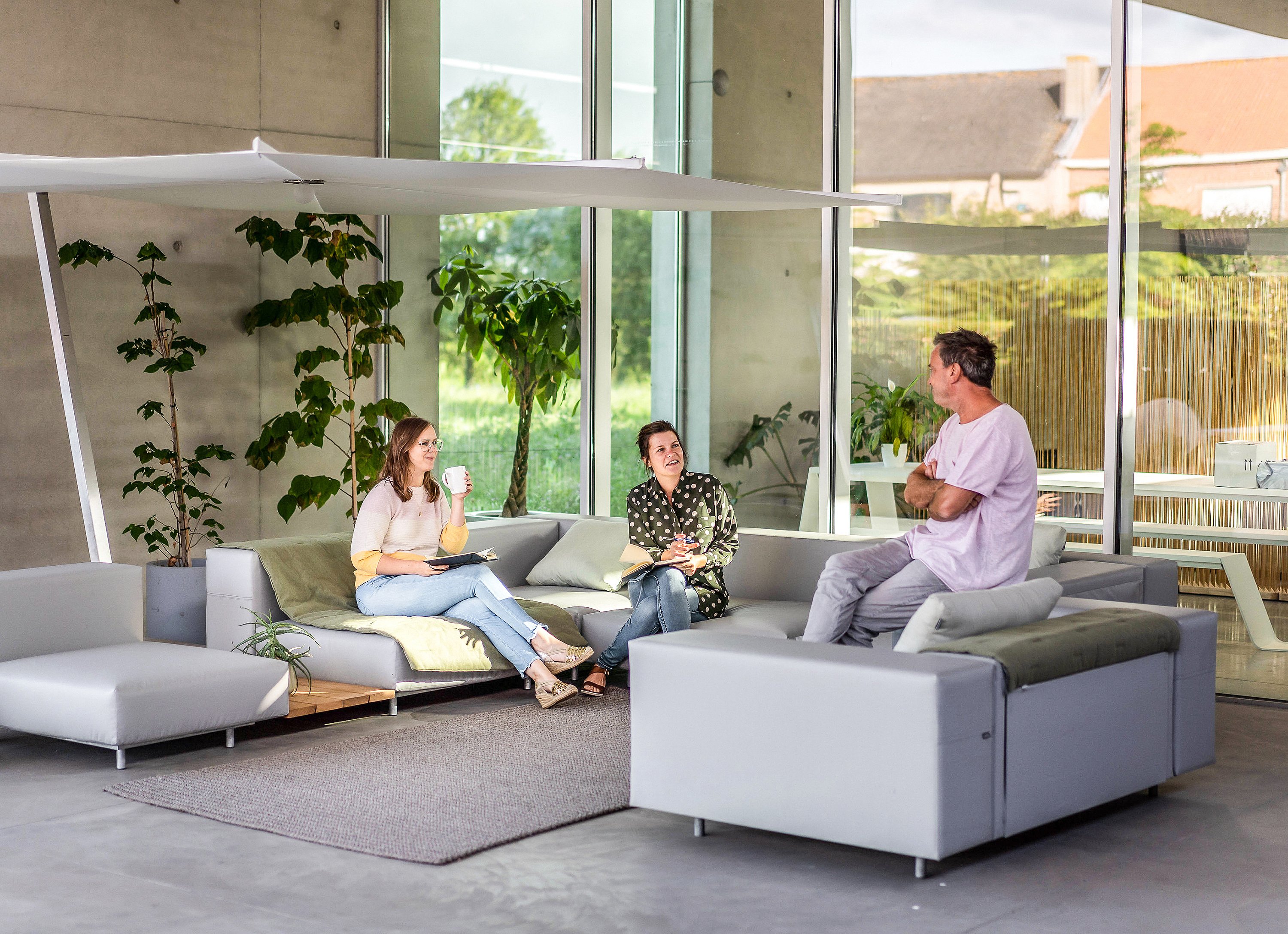   Drei Kollegen entspannen und unterhalten sich auf modularen Gartensofas in einer überdachten Terrassenlounge mit hohen Fenstern und viel Grün.