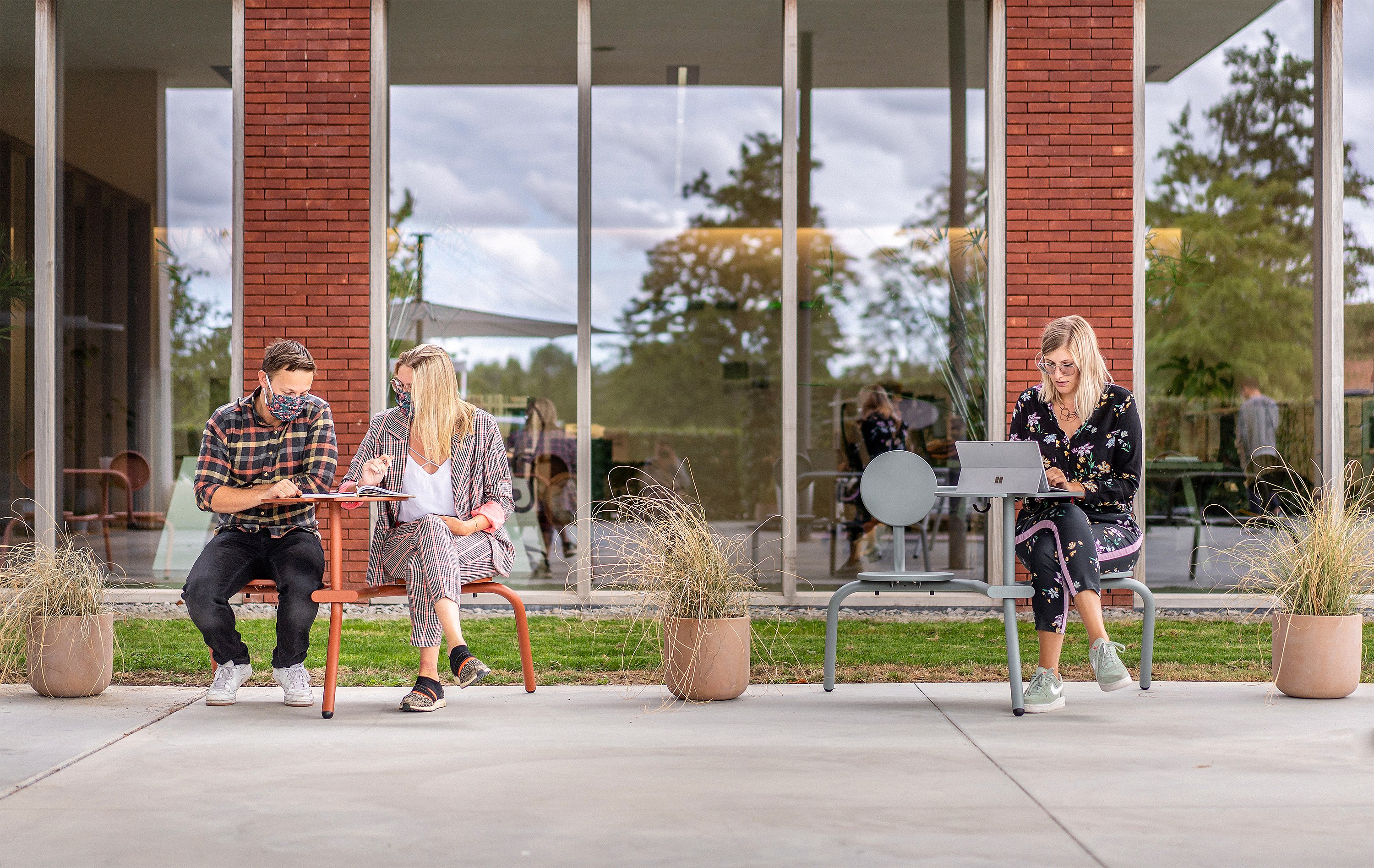 Trois collègues travaillant à l’extérieur sur des sièges et des tables de style bistroo compacts devant un bâtiment de bureaux en brique et verre.