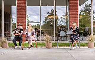 Trois collègues travaillant à l’extérieur sur des sièges et des tables de style bistroo compacts devant un bâtiment de bureaux en brique et verre.
