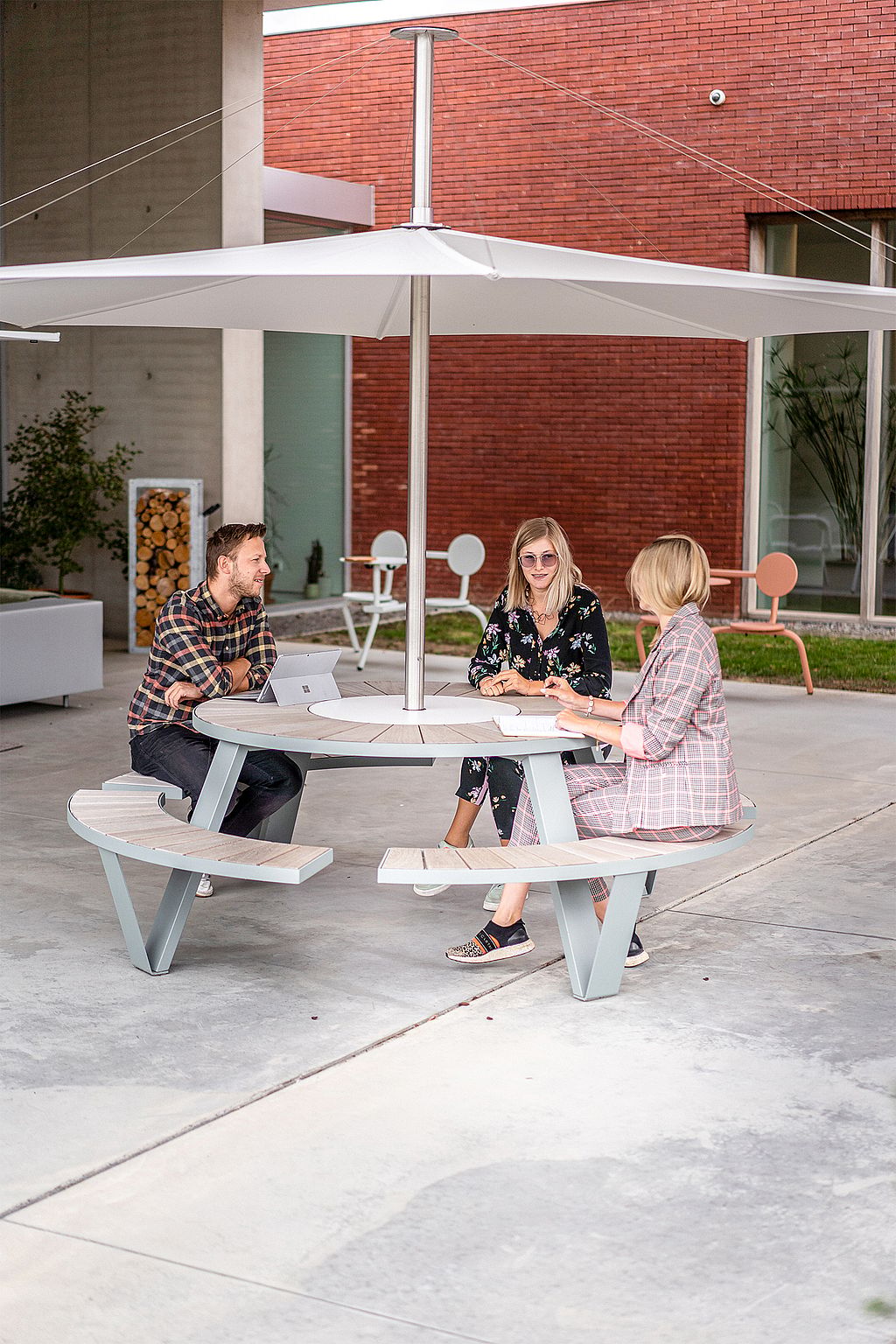 Three colleagues seated around a round outdoor table with a central parasol, talking and working in a modern courtyard with brick walls behind them.