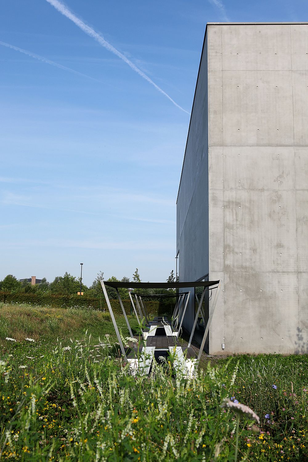 Longue table de pique-nique Hopper ombragée longe un grand bâtiment en béton, entourée d'une prairie naturelle.