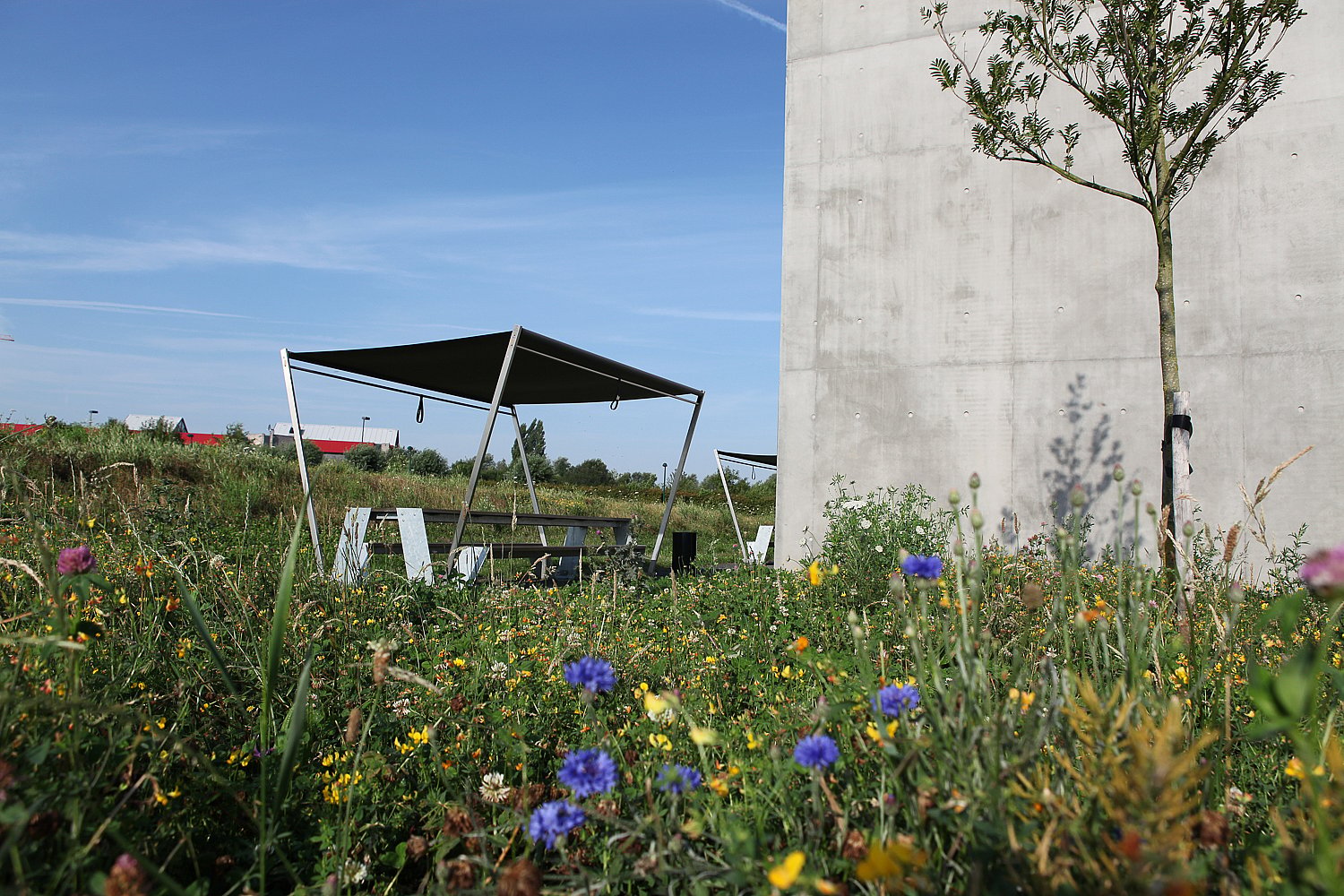 Hopper picnic table with a dark shade set in a wildflower meadow beside a tall concrete building, with open sky above.