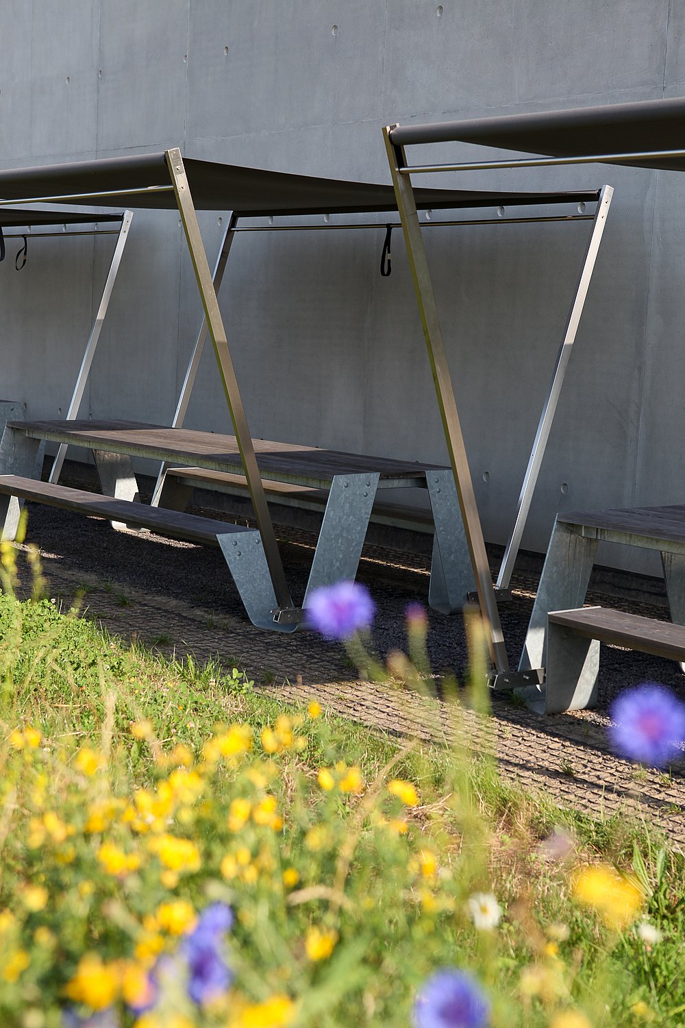   Gros plan sur des tables de pique-nique Hopper avec parasols sombres et assises en bois, près d'un mur en béton, avec des fleurs sauvages au premier plan.