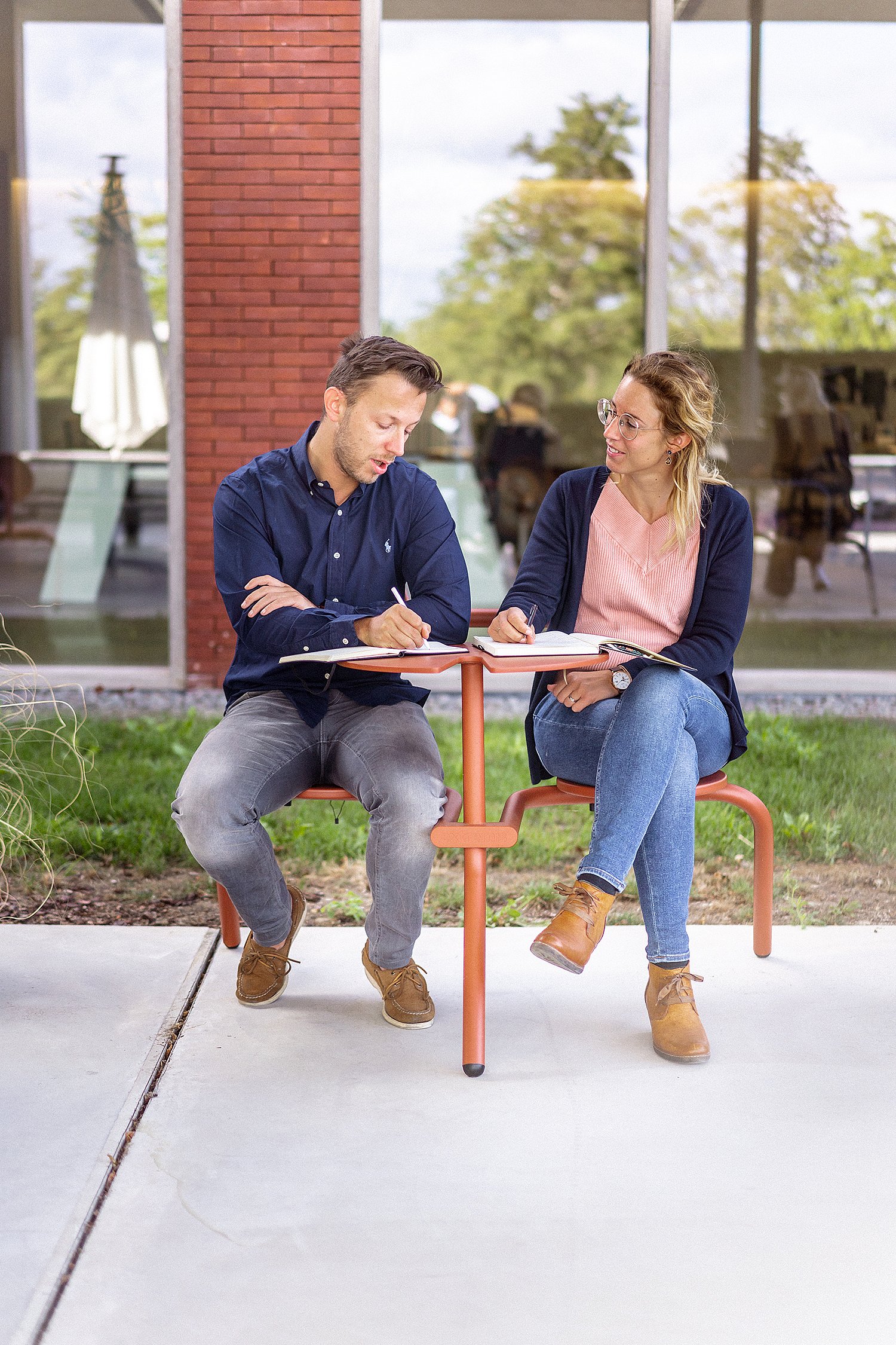 Deux collègues écrivent dans des cahiers tout en étant assis à une petite table bistroo rouillée.