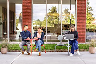 Trois collègues travaillant à l’extérieur sur des sièges et tables compacts de style bistro devant un immeuble de bureaux en brique et verre.