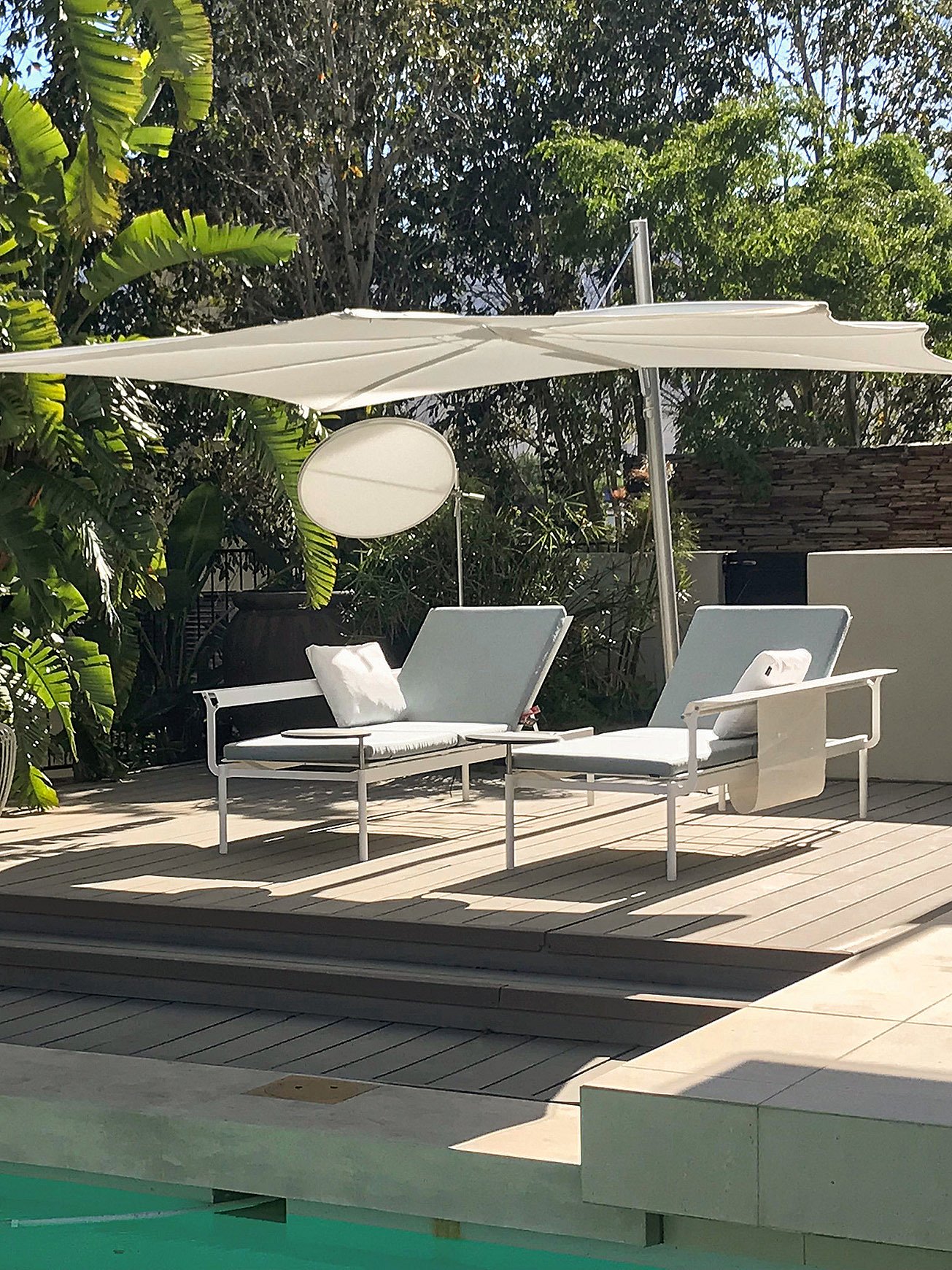 Zwei weiße Sol+Luna-Liegen mit gemeinsamem Schatten in der Nabelschnur auf einem Pooldeck aus Holz, umgeben von üppiger tropischer Bepflanzung.
