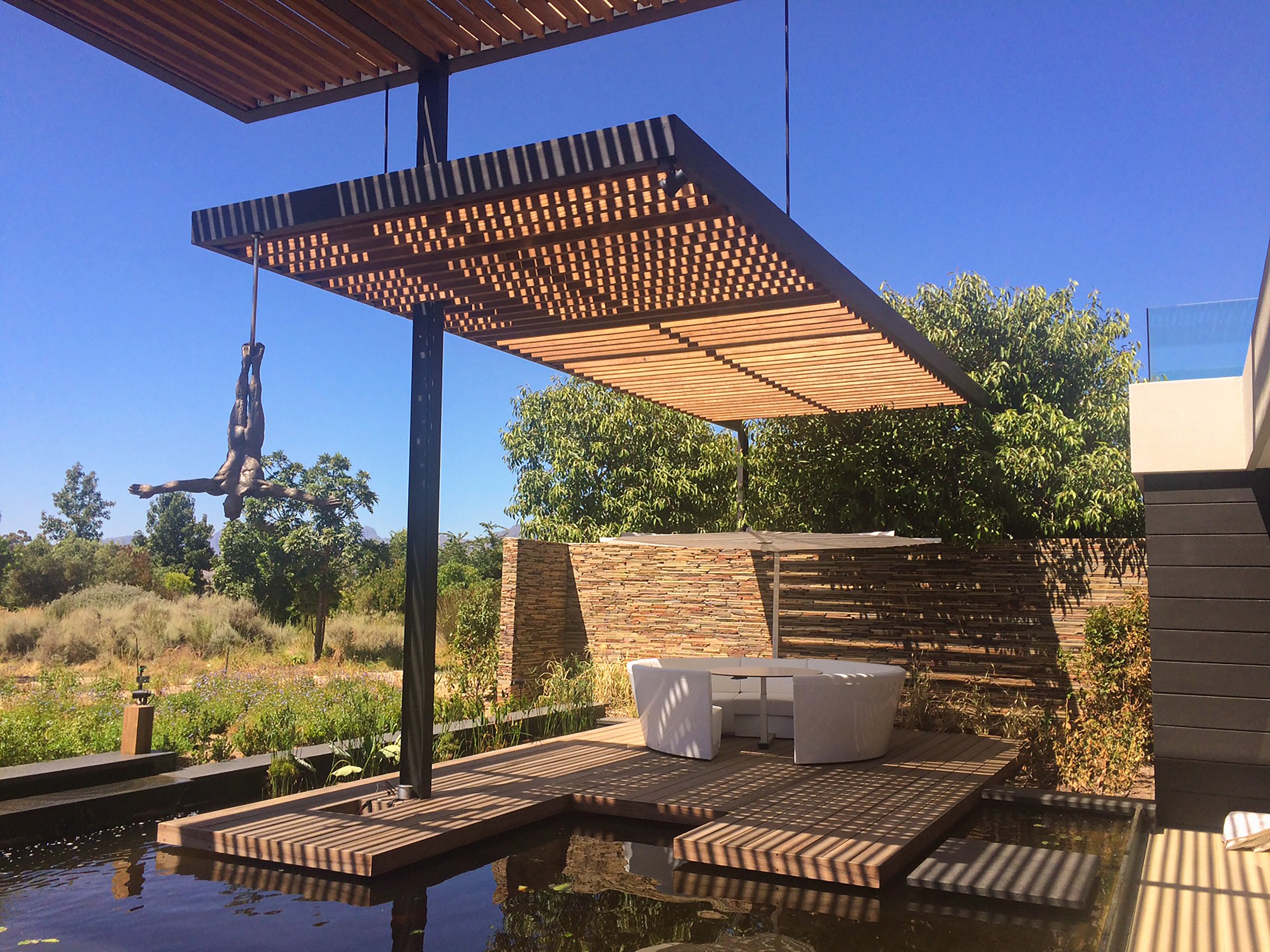 Kosmos Outdoor-Lounge unter einem Lattendach neben einem reflektierenden Pool, mit Bäumen und steinernen Gartenmauern im Hintergrund.