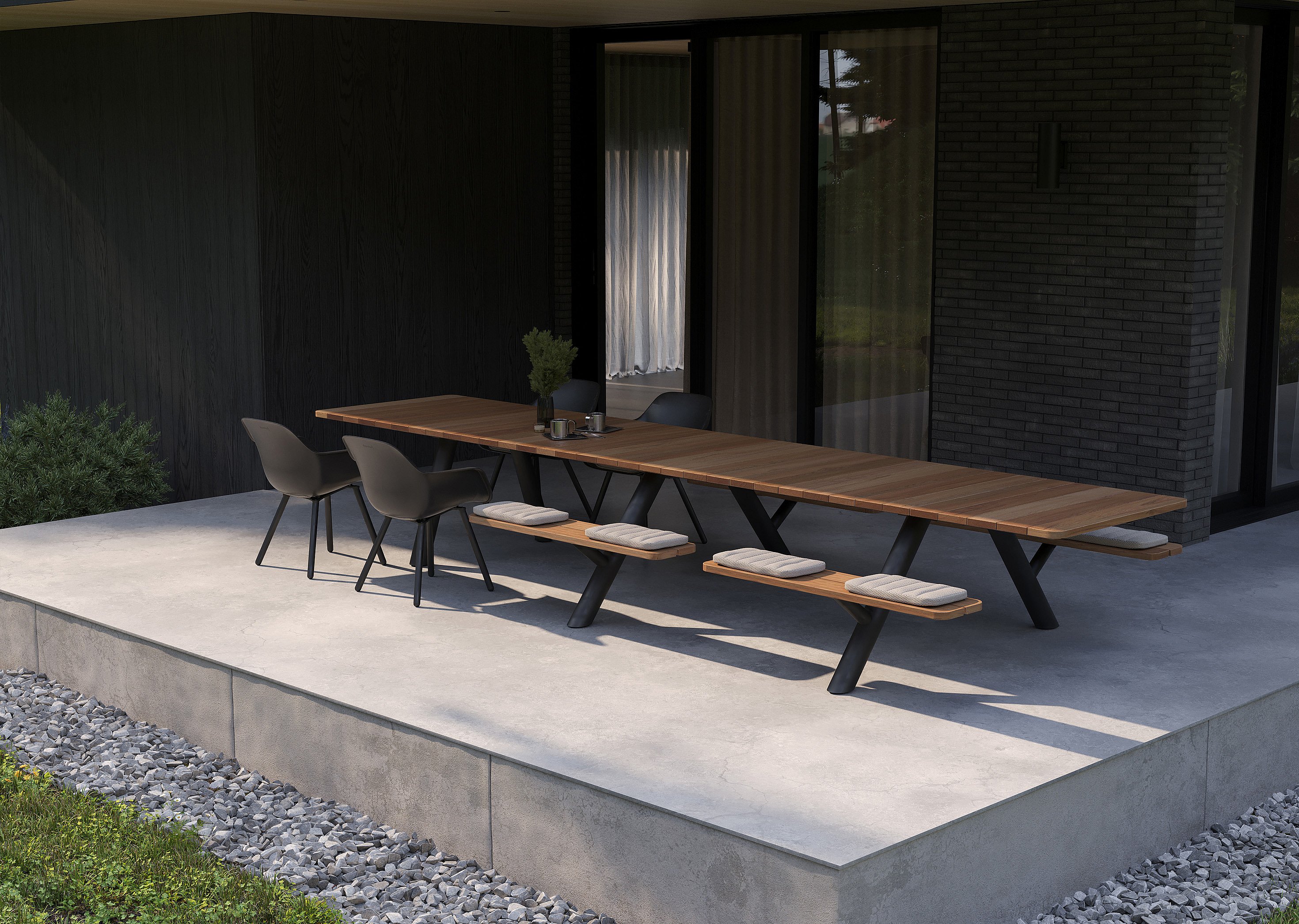 Angled view of the long Panigiri table with bench cushions and black chairs on a minimalist concrete terrace beside the house.
