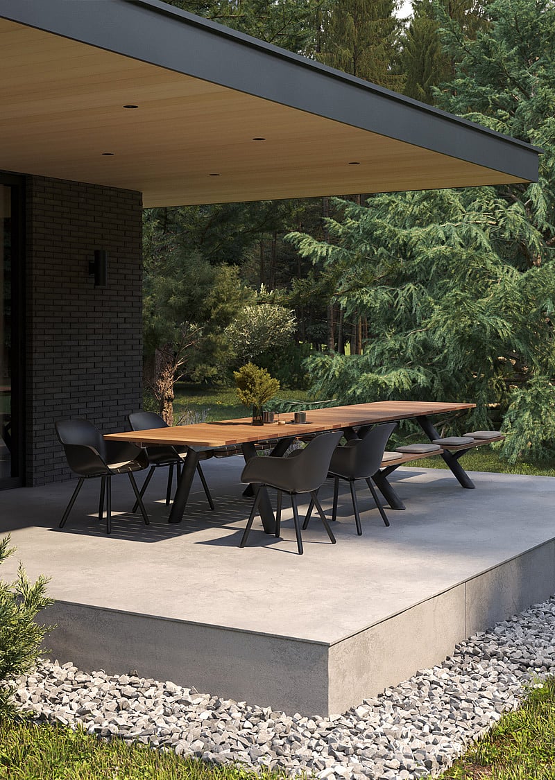Long wooden Panigiri table with benches and black chairs on a raised terrace under a deep roof overhang, with a pine forest in the background.