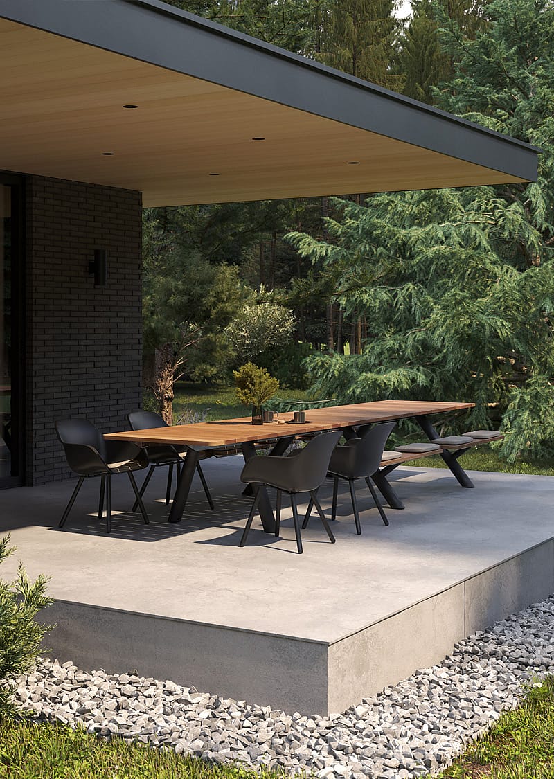 Long wooden Panigiri table with benches and black chairs on a raised terrace under a deep roof overhang, with a pine forest in the background.