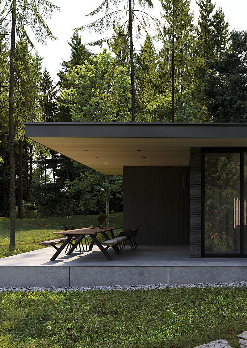 Side view of a modern bungalow terrace with a long Panigiri table tucked beneath a broad roof, surrounded by lawn and tall pine trees.