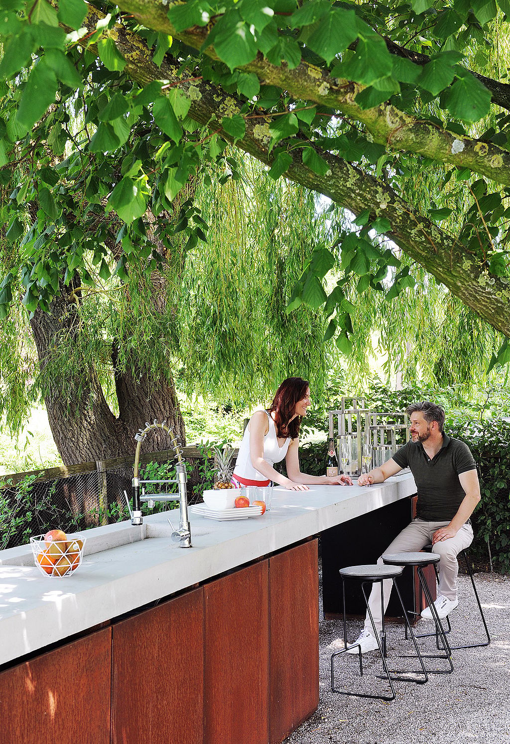 A woman leaning on a long outdoor kitchen counter while talking to a seated man on a black Tiki barstool, framed by overhanging green branches.