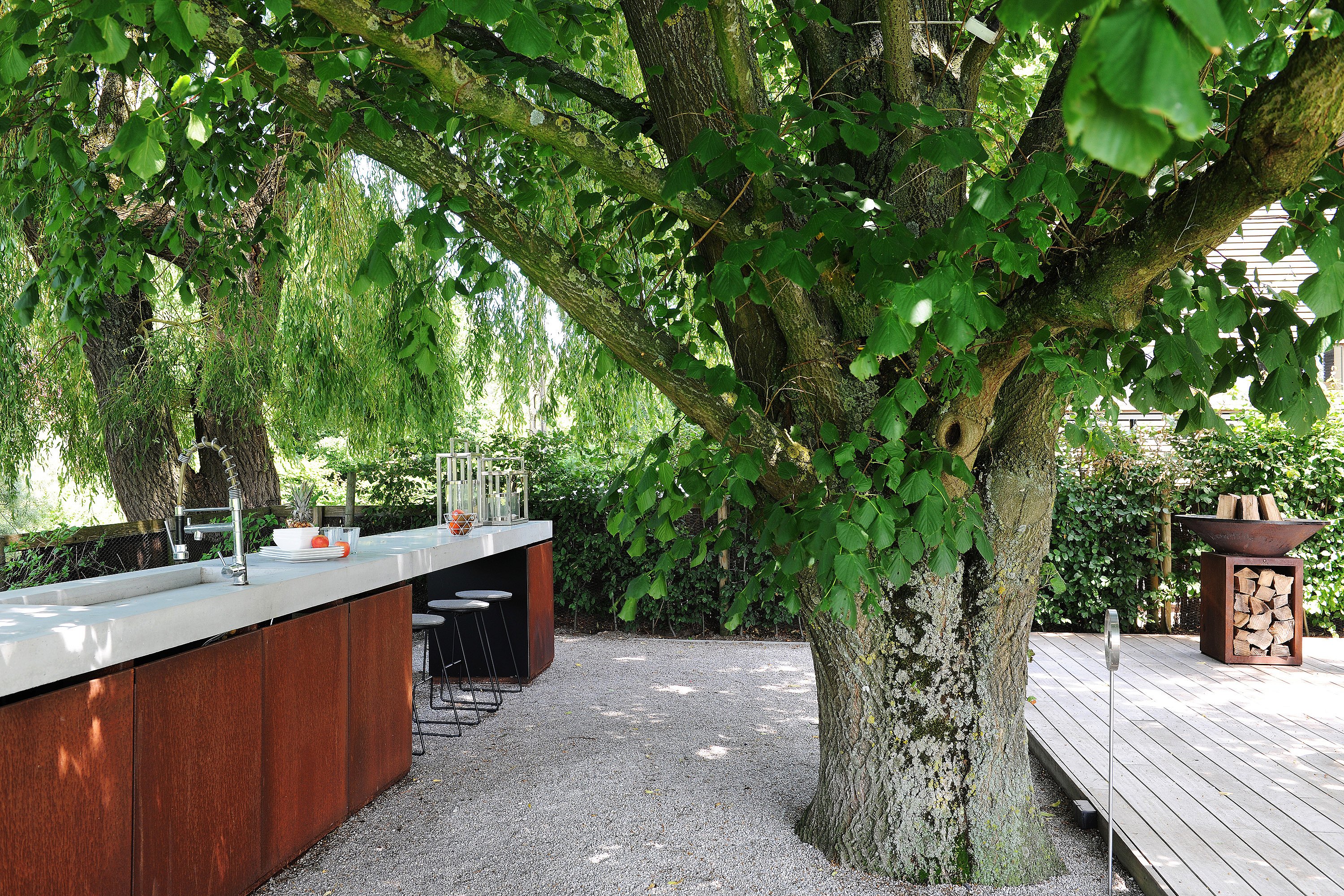 Een roestkleurig buitenkeukeneiland met een wit aanrechtblad en zwarte Tiki-barkrukken onder een grote, lommerrijke boom, naast een houten terras met een vuurkorf.
