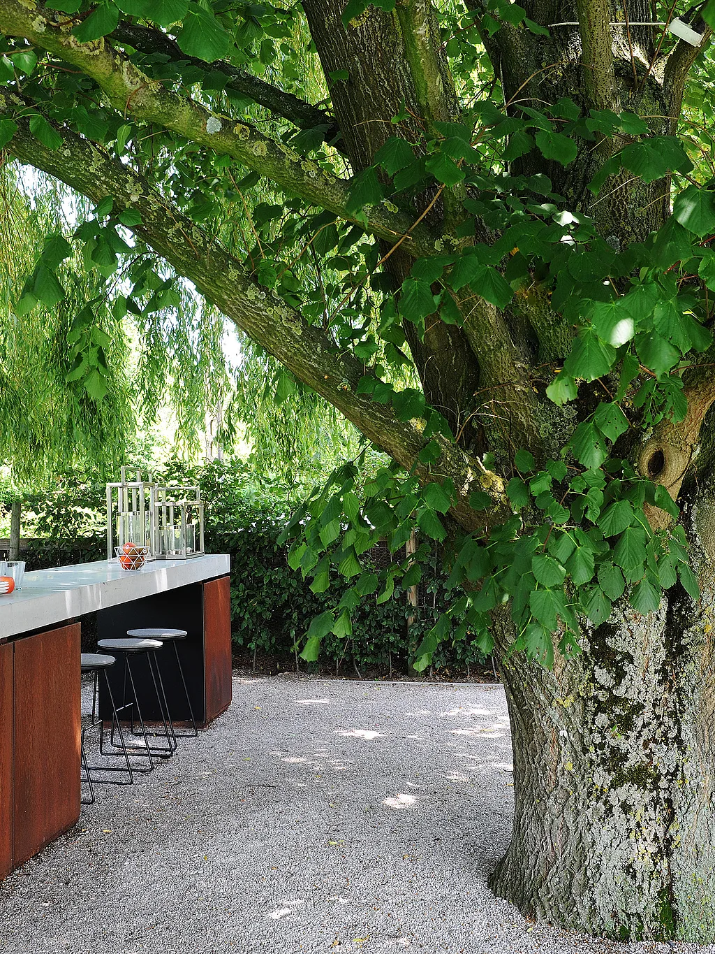 Een roestkleurig buitenkeukeneiland met een wit aanrechtblad en zwarte Tiki-barkrukken onder een grote, lommerrijke boom, naast een houten terras met een vuurkorf.