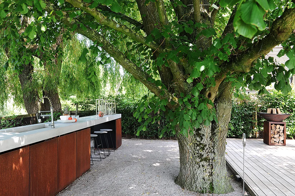 Rostfarbene Kücheninsel im Freien mit weißer Arbeitsplatte und schwarzen Tiki-Barhockern unter einem großen, belaubten Baum, neben einer Holzterrasse mit Feuerschale.