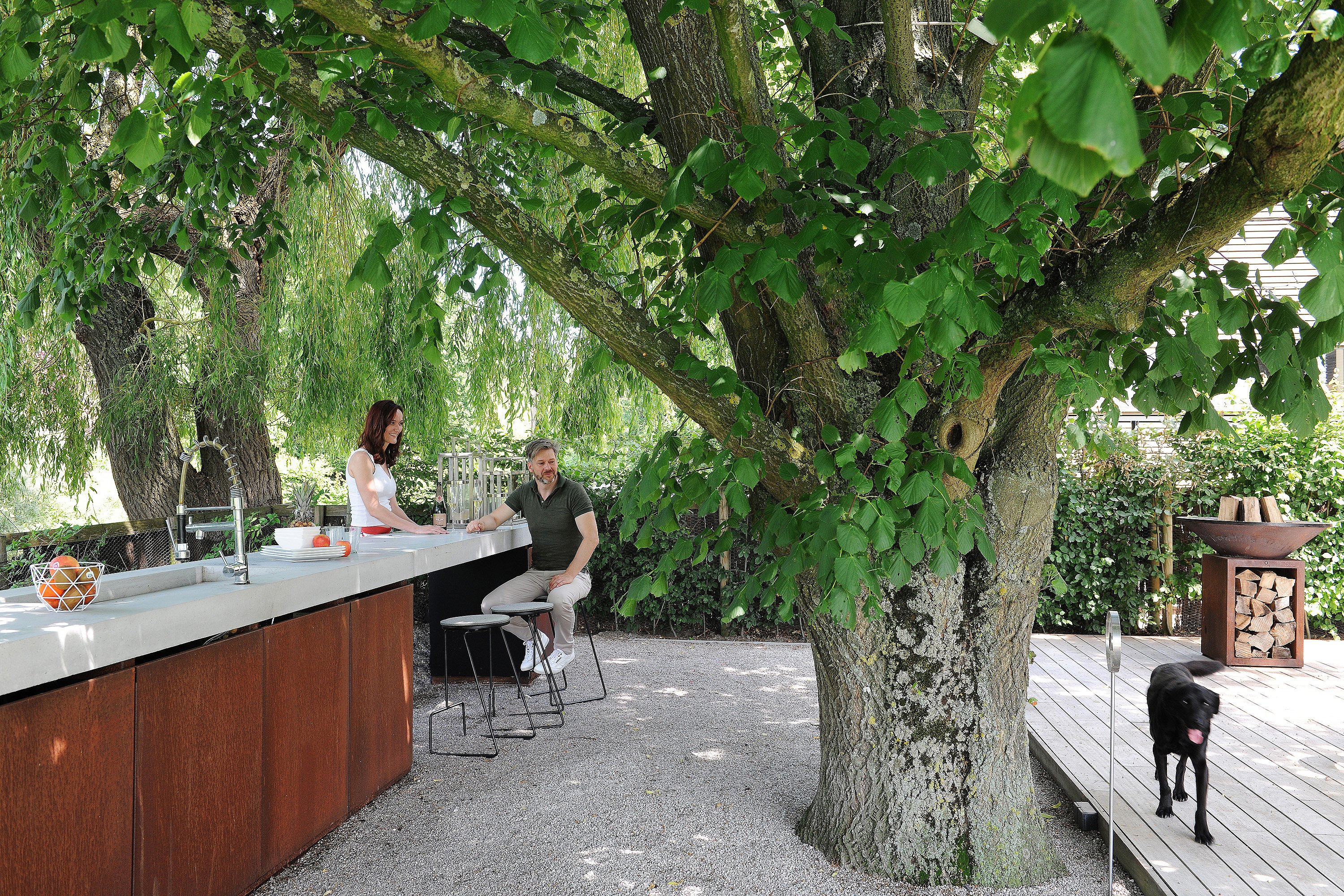 A woman and a man at a long outdoor kitchen counter under a large tree, with black Tiki barstools, gravel ground, and a black dog walking on the deck nearby.