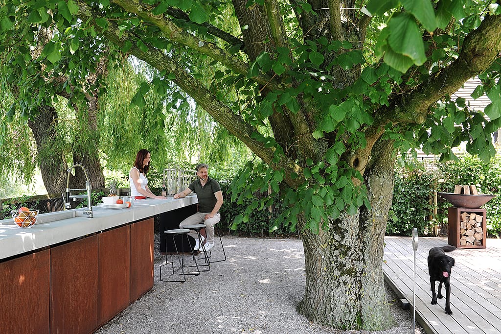 A woman and a man at a long outdoor kitchen counter under a large tree, with black Tiki barstools, gravel ground, and a black dog walking on the deck nearby.