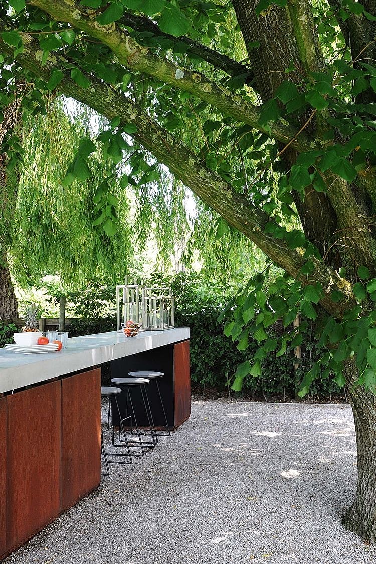 Close view of the outdoor kitchen island with a white countertop, black Tiki barstools, and decorative lanterns beneath a large tree in a shaded garden.
