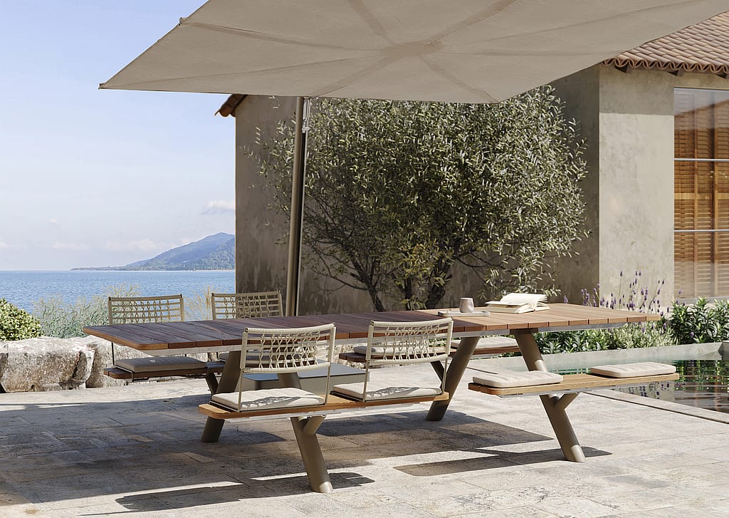 Wooden Panigiri picnic table with woven BYOS backrests and seat cushions under a large Sensu shade, overlooking the sea beside a villa terrace.