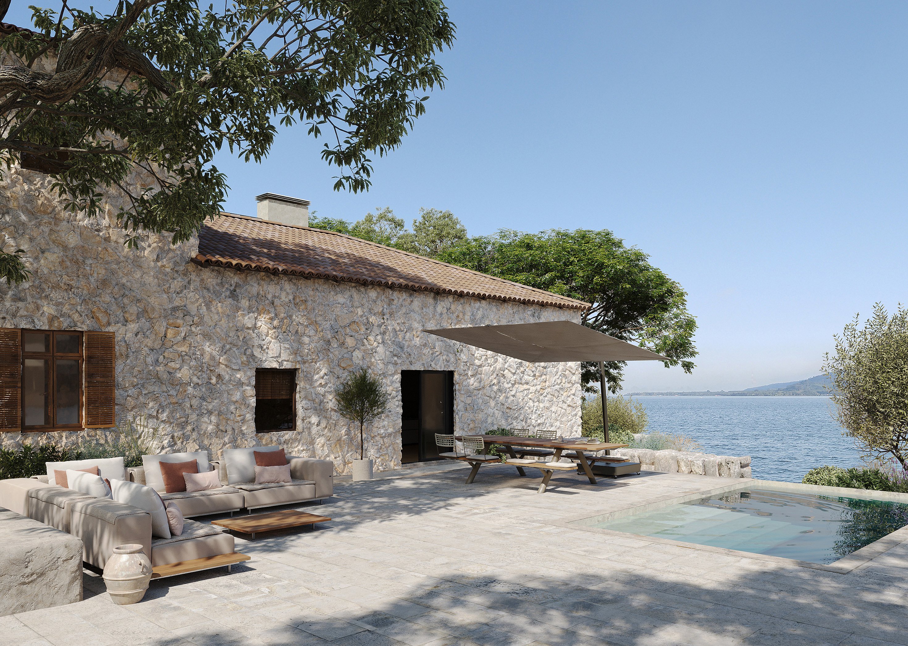 Beige and terracotta outdoor walrus seating and a Panigiri picnic table with shade on a stone terrace beside the sea, outside a rustic Mediterranean villa.