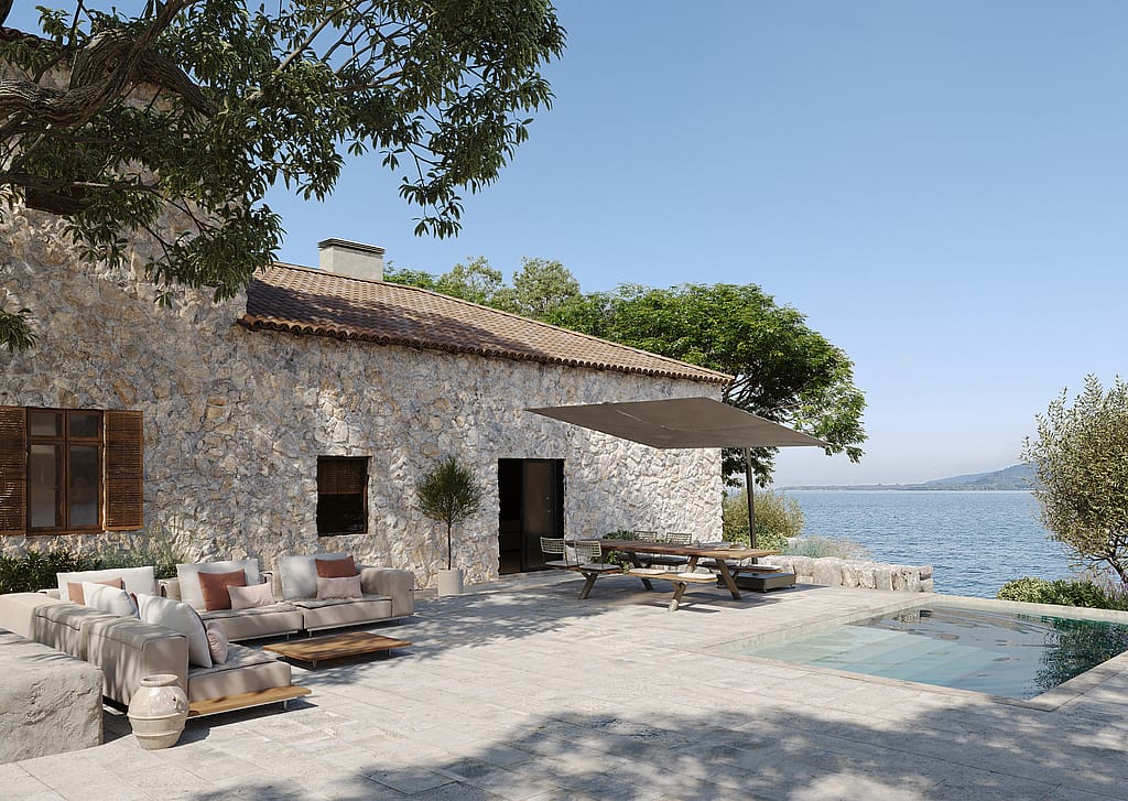 Beige and terracotta outdoor walrus seating and a Panigiri picnic table with shade on a stone terrace beside the sea, outside a rustic Mediterranean villa.