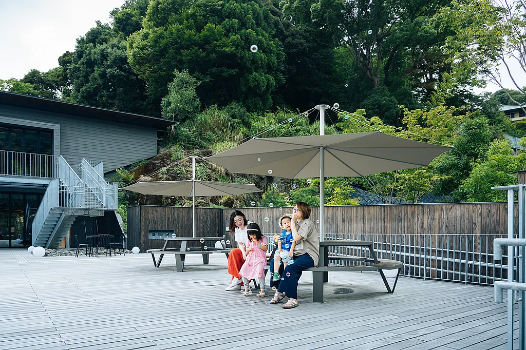 Families sitting at round Pantagruel picnic tables under large Inumbra parasols on a spacious wooden terrace surrounded by trees.