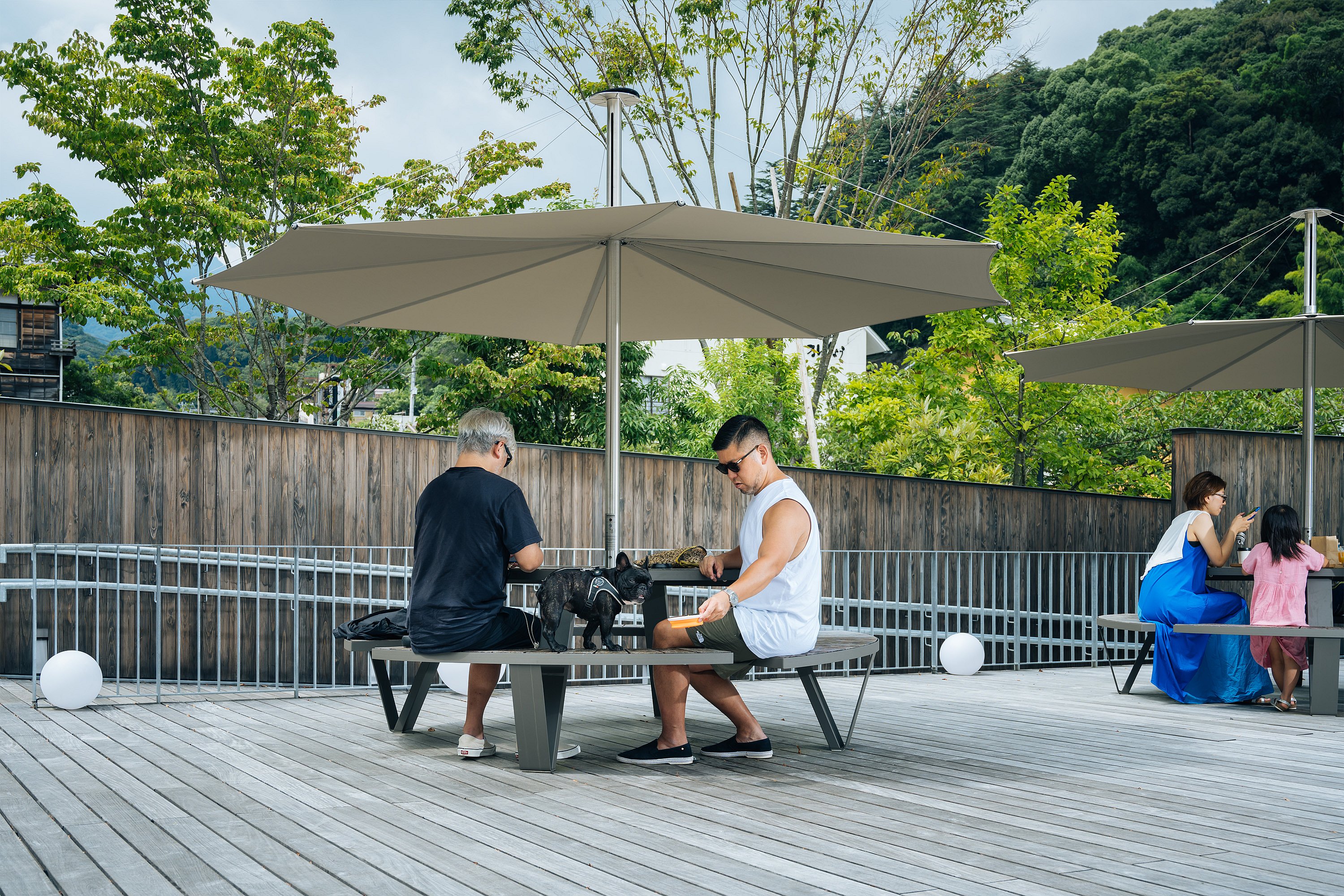 Deux personnes assises à une table de pique-nique ronde Pantagruel sous un parasol Inumbra sur une terrasse en bois, avec un autre repas familial en arrière-plan.