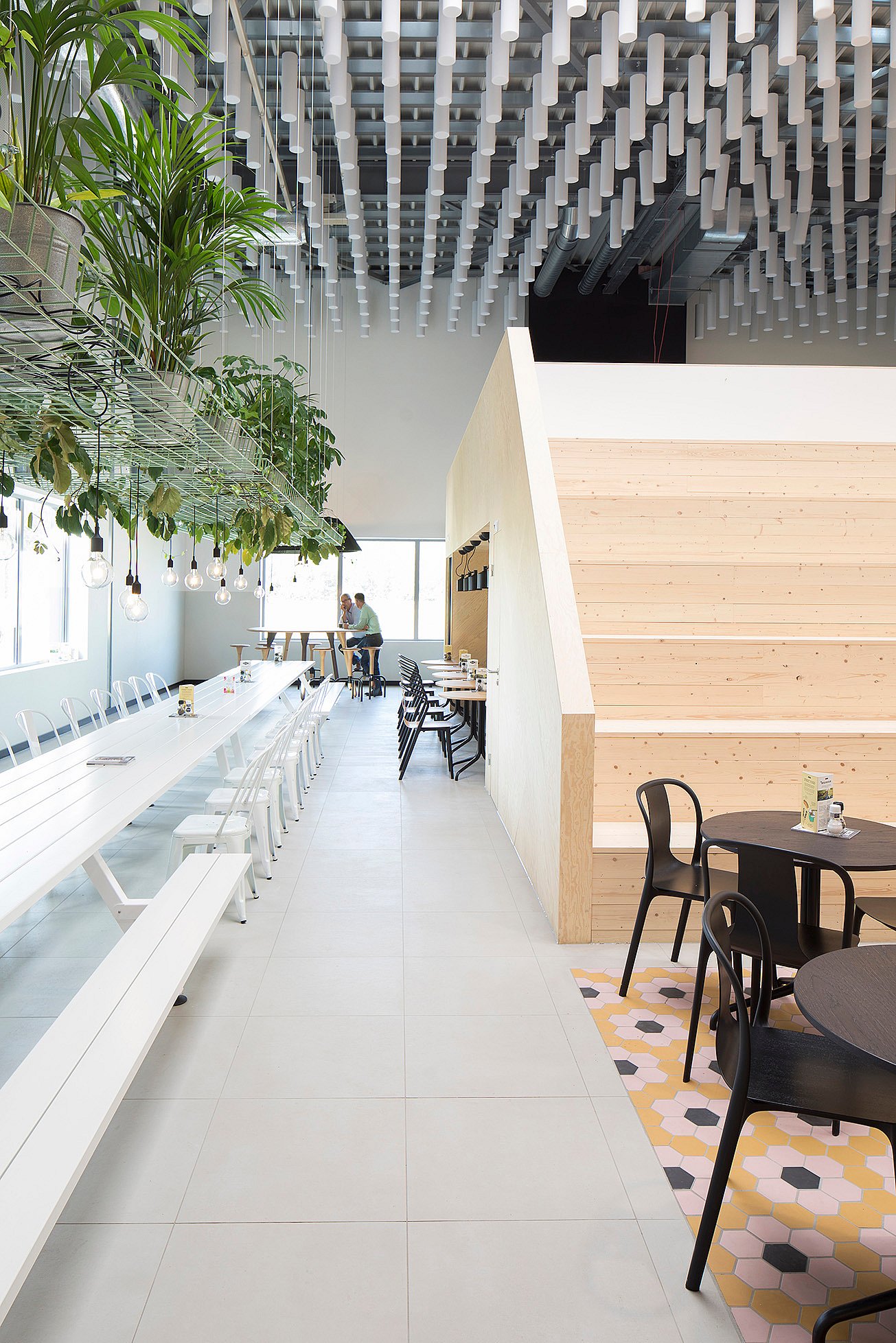 Long white communal Marina- lunch table with benches in a bright modern canteen, beside hanging plants, pendant lights, black café tables, and wide wooden steps.