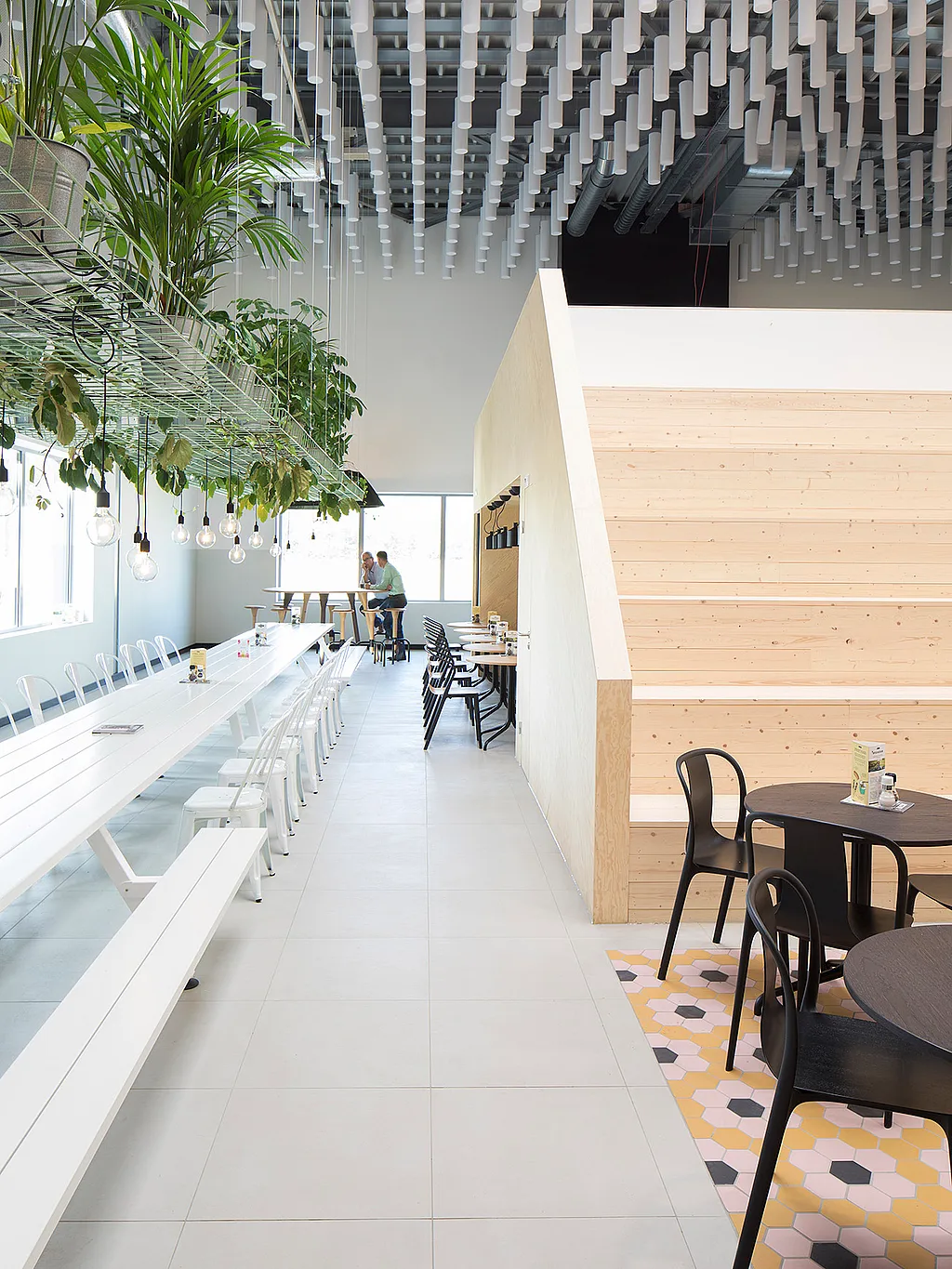 Long white communal Marina- lunch table with benches in a bright modern canteen, beside hanging plants, pendant lights, black café tables, and wide wooden steps.