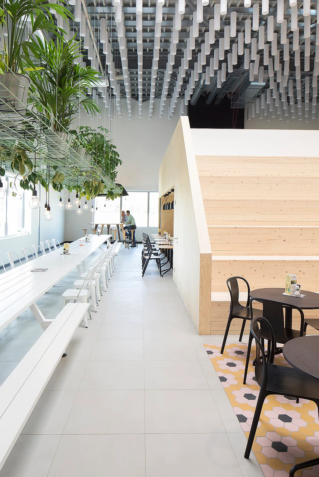 Lange witte Marina-lunchtafel met banken in een lichte, moderne kantine, naast hangplanten, pendellampen, zwarte cafétafels en brede houten trappen.