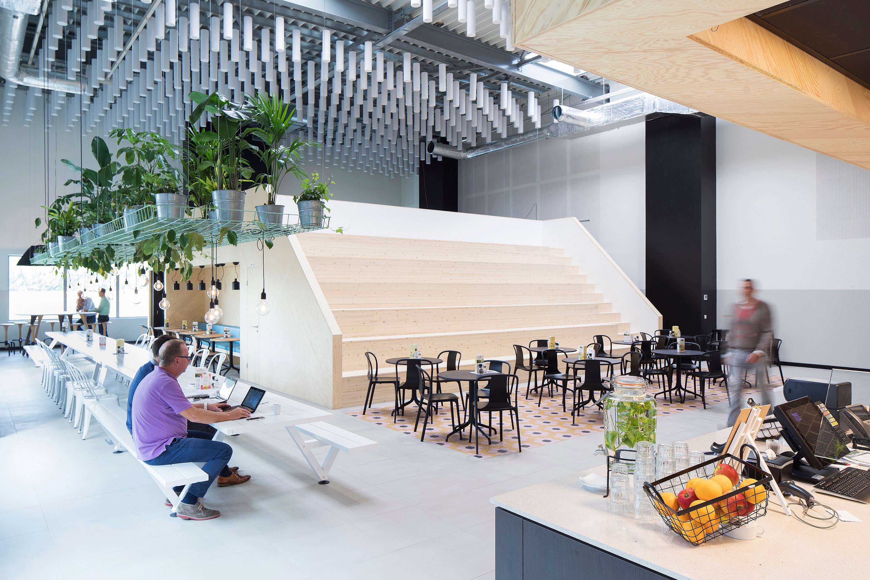 Breed zicht op een moderne fabriekskantine met een lange witte Marina-tafel, cafézitjes op tegels met een patroon, hangplanten en brede houten zittreden.