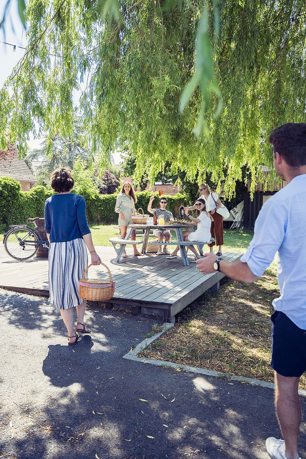 Freunde mit Picknickkörben gehen unter einer großen Weide auf den Gargantua-Tisch zu, während andere warten und vom Tisch winken.