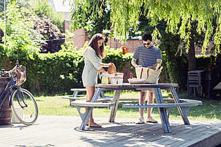 Zwei Personen packen einen Picknickkorb aus und arrangieren Essen und Getränke auf einem runden Gargantua-Picknicktisch auf einem Holzdeck, neben dem ein Fahrrad geparkt ist.