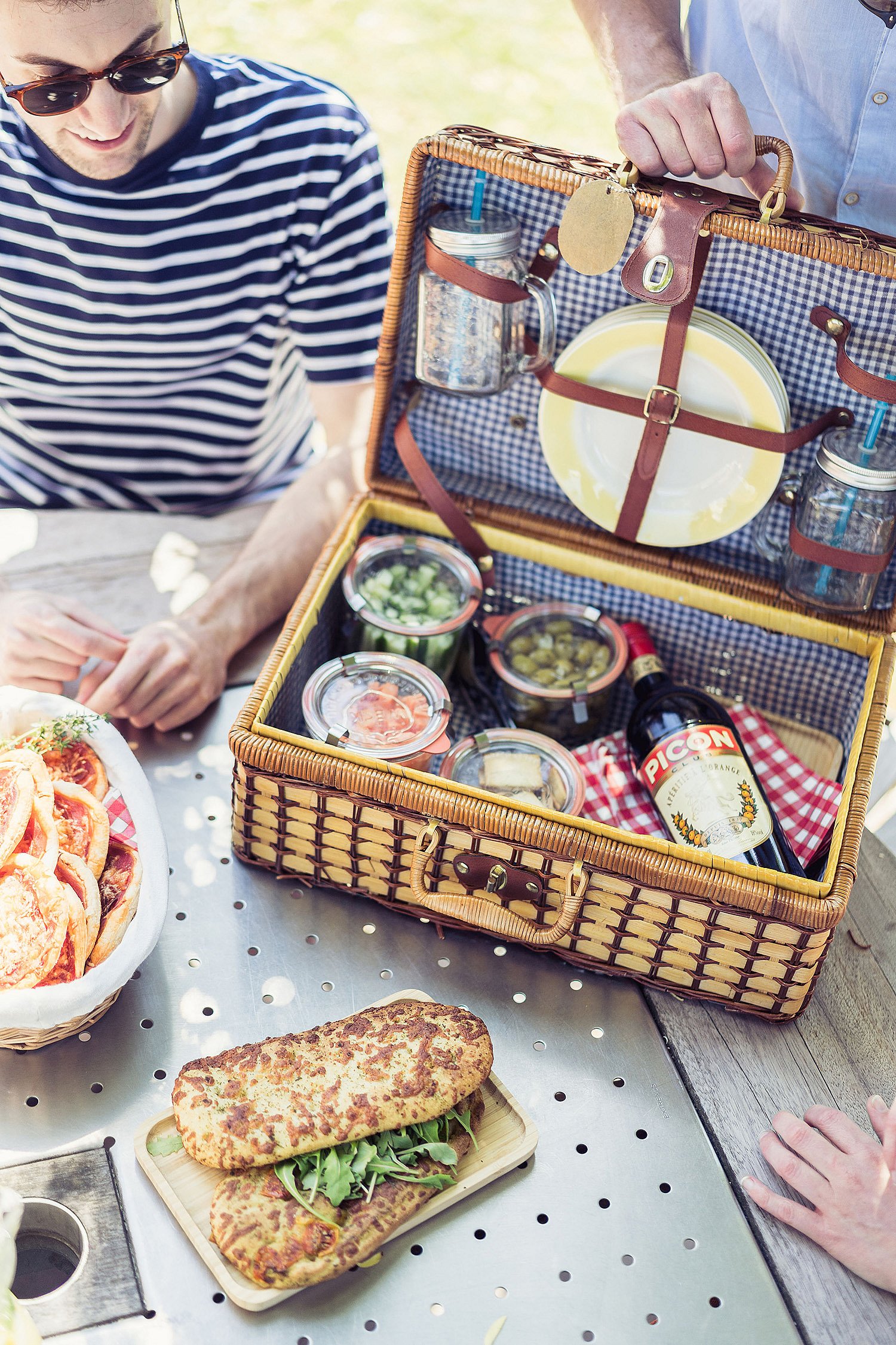 Open wicker picnic basket filled with plates, glasses, jars and drinks on a picnic table beside sandwiches and other shared food.