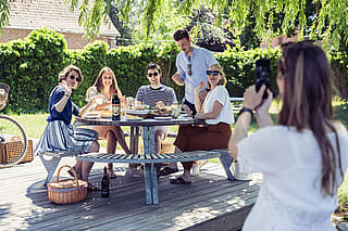 Group portrait of five friends smiling and waving at the camera around a round outdoor picnic table in a sunny garden.