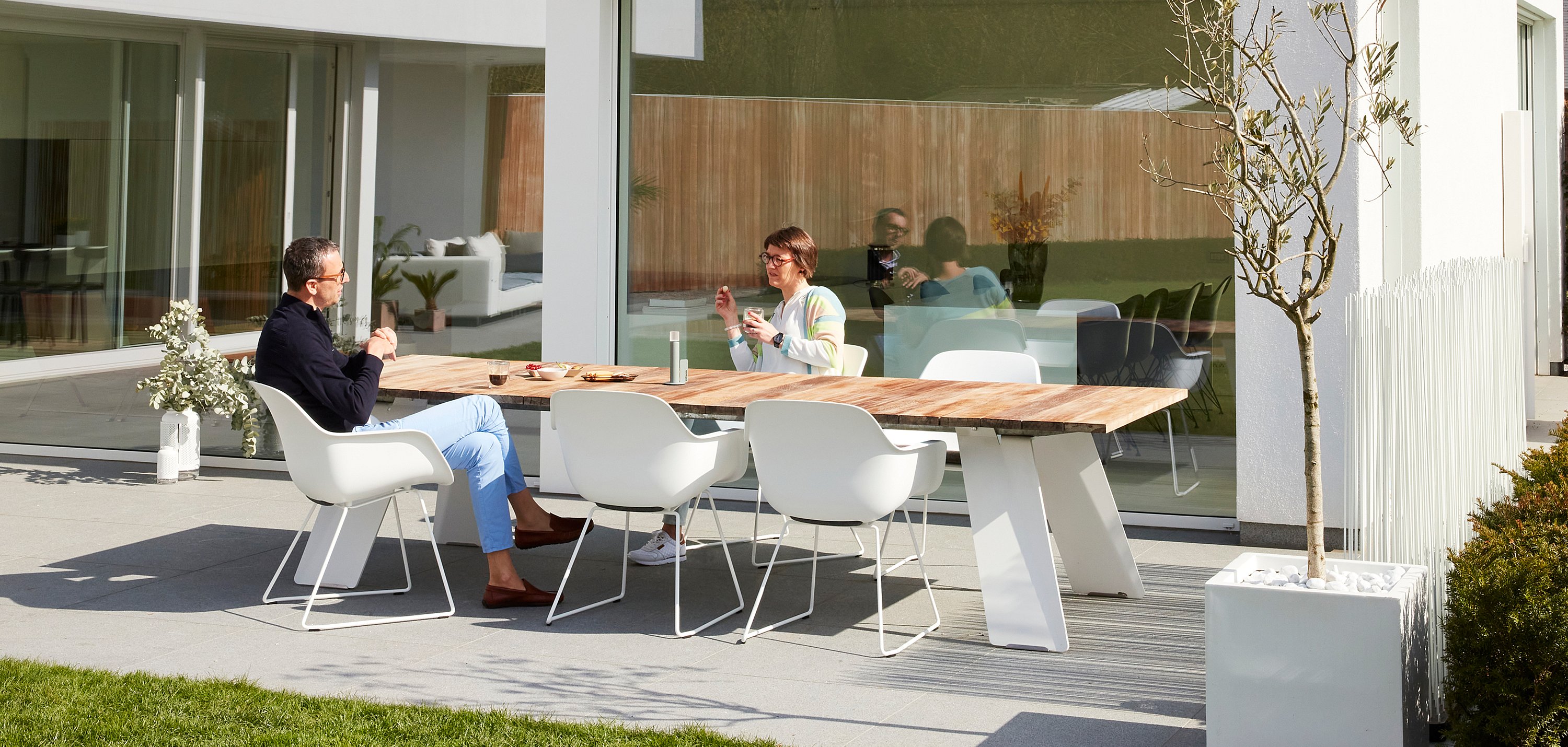 Menschen sitzen um einen Tisch im Freien und genießen Kaffee und Gebäck in einem sonnigen Hinterhof, der zum Entspannen und geselligen Beisammensein gedacht ist.
