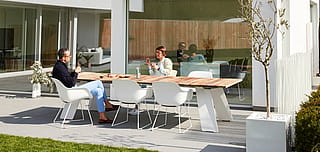 Menschen sitzen um einen Tisch im Freien und genießen Kaffee und Gebäck in einem sonnigen Hinterhof, der zum Entspannen und geselligen Beisammensein gedacht ist.