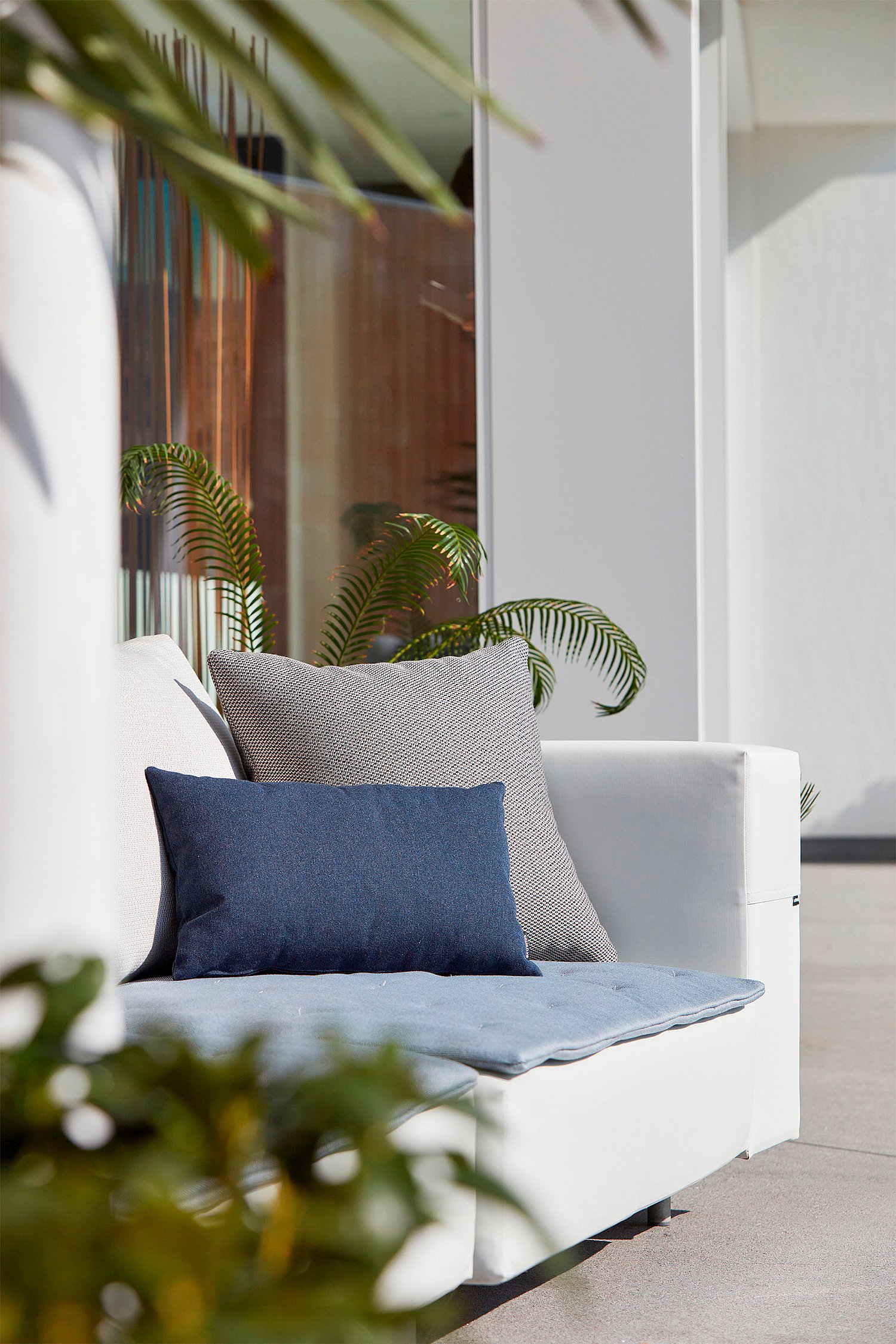 Weiche Kissen in hellen Tönen, die auf einem weißen Outdoor-Sofa angeordnet sind und Textur und Komfort in einer raffinierten Gartenumgebung hervorheben.